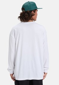 Langärmliges weißes T-Shirt, runder Halsausschnitt, überschnittene Schultern, weiches Baumwollmaterial, schlichte Textur, präsentiert an einer Person, die eine grüne Mütze trägt.