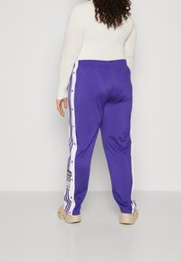 Fialové teplákové kalhoty s bílými bočními pruhy a cvoky, vyrobené z měkkého materiálu a s elastickým pasem. Etiketa viditelná poblíž lemu.