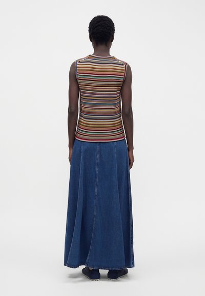 Personne de dos portant un top sans manches à rayures multicolores, une longue jupe en jean bleu foncé et des chaussures noires.