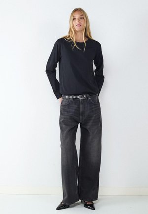 Jeune femme portant un haut noir à manches longues, un jean gris foncé à jambes larges, une ceinture cloutée et des chaussures noires pointues, debout contre un mur blanc.