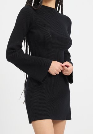 Frau trägt ein schwarzes, geripptes Strick-Minikleid mit ausgestellten Ärmeln und steht mit vor dem Körper verschränkten Händen.