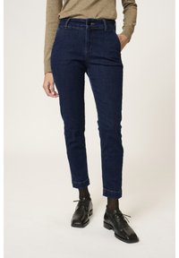 Jean bleu foncé à coupe slim, en denim. Comprend une taille haute, des poches avant et un ourlet retroussé. Associé à des chaussures noires à lacets.