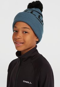 Cappello di lana blu con un pompon nero e testo nero a contrasto. Chi lo indossa ha una giacca nera con zip che presenta un logo bianco.