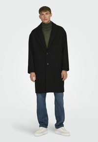 Abrigo de mezcla de lana negro con cuello con muescas, botones frontales y bolsillos laterales, llevado sobre un cuello de tortuga de punto verde y jeans azules. Zapatillas blancas.