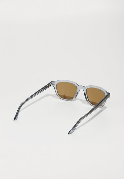 Graue, transparente Cat-Eye-Sonnenbrille mit braun getönten Gläsern. Schlanke Bügel mit subtilen Branding-Details. Minimalistisches Design, glatte Textur.