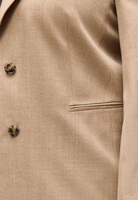 Κοντινό πλάνο υφάσματος εκρού σακακιού με δύο καφέ κουμπιά και μια οριζόντια τσέπη με σχισμή στη δεξιά πλευρά.