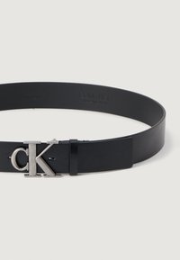 Zwarte leren riem met een gladde textuur, voorzien van een zilveren metalen "CK" logo bevestiging en een minimalistisch ontwerp. Verstelbaar met een klassieke sluiting.