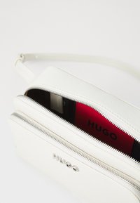 Witte handtas met een gepolijste textuur, voorzien van twee met rits afgesloten compartimenten en een rode binnenkant. Zilveren logo aan de voorkant. Verstelbare schouderband inbegrepen.