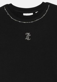 Melna t-kreklā ar apaļu kakla izgriezumu, rotāta ar sudraba pērlītēm ap kaklu un mazu pērlīšu logotipu uz krūšu.