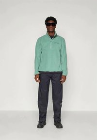 Ανοιχτό πράσινος φλις πόλο με μισό φερμουάρ και λογότυπο στο αριστερό στήθος, φορεμένος με σκούρους παντελόνες και μαύρα παπούτσια από μοντέλο.