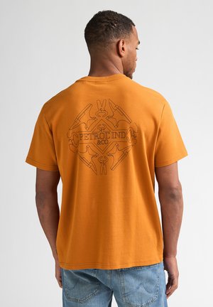 Man draagt een oranje T-shirt met een zwart geometrisch "PETROL IND & CO" logo op de rug, gecombineerd met lichtblauwe spijkerbroek, staand tegen een effen achtergrond.