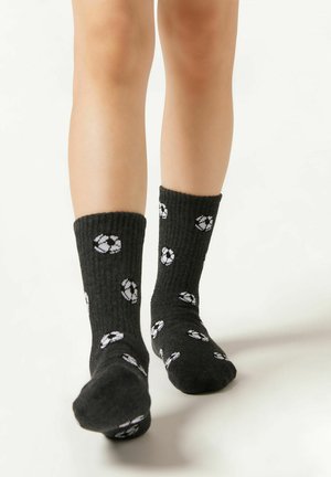 Svarta ribbade strumpor med vita återvinningssymboler, med en höjd till mitten av vaden och mjuk textur, visade på ett barns fötter.