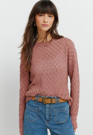 Jonge vrouw met lang bruin haar, gekleed in een roze gebreide trui en een blauwe spijkerbroek met voorzakken en een beige riem