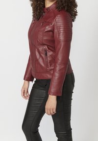 Czerwona skórzana kurtka z pikowanym wykończeniem na ramionach, zamkami z przodu i dopasowanym krojem. W zestawieniu z czarnymi, teksturowanymi legginsami.