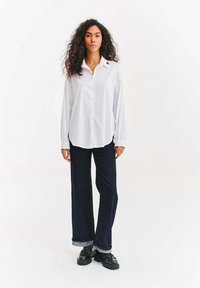 Biała, długorękawowa koszula na guziki z zaokrąglonym dołem i małym logo na kołnierzyku, zestawiona z ciemnymi, szerokimi dżinsami i czarnymi butami.