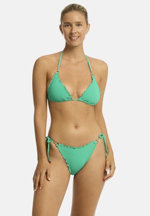 Kobieta w teksturowanym turkusowym bikini z wiązanymi ramiączkami, uśmiechająca się, stojąca na jednolitym jasnym tle.