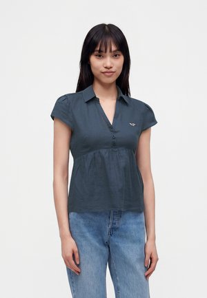 Junge Frau mit glatten schwarzen Haaren, die eine dunkelblaue kurzärmelige Bluse mit Knöpfen und hellblaue Jeans trägt und vor einem unifarbenen Hintergrund steht.