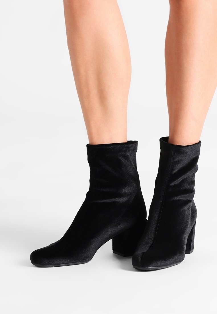 Bottines en velours noir à talons blocs portées sur jambes nues sur fond blanc.