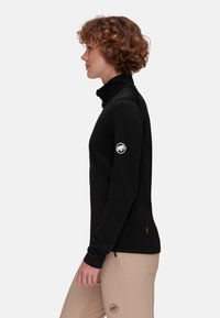 Giacca in pile nera con colletto alto, chiusura con zip e logo sul braccio sinistro. Texture liscia e design aderente, abbinata a pantaloni beige.