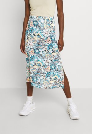 Femme portant une jupe mi-longue fleurie avec des motifs bleus, pêche et noirs, associée à des baskets blanches et des chaussettes, debout les jambes écartées.