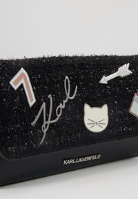 Borsa nera texture con hardware argento, caratterizzata da un motivo con firma e gattino, e vari spilloni in smalto colorato. Base in pelle liscia.