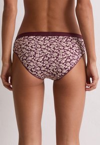 Femme de dos, portant des sous-vêtements violets à motifs floraux blancs, se tenant devant un mur clair.