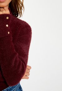 Donna che indossa un maglione fuzzy bordeaux con bottoni dorati sui polsini, toccandosi il viso, e jeans in denim blu su uno sfondo bianco.