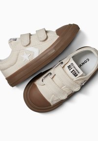 Zapatillas de lona beige con dos correas de Velcro, puntera y suela de goma marrón, que presentan un logo de estrella blanca y costuras en contraste.