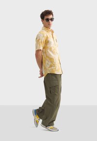 Korte mouwen gele shirt met witte bladvormen, gecombineerd met olijfgroene cargobroeken en multicolore sneakers. Model draagt zwarte zonnebril.