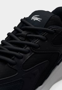 Κοντινό πλάνο μαύρου αθλητικού παπουτσιού με υφασμάτινα πάνελ, στρογγυλά μαύρα κορδόνια και λευκό λογότυπο κροκοδείλου στη γλώσσα πάνω από λευκή σόλα.