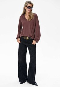 Vrouw in oversized zonnebril, bruine blouse met lange mouwen, zwarte wijde broek, zwarte riem met gouden gesp en zwarte puntige schoenen.
