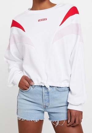 Femme portant un sweat-shirt blanc Levi's avec des rayures en chevron rouges et roses et un short en denim bleu clair effiloché, main dans la poche.