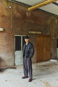 Junger Mann trägt schwarze Strickmütze, schwarze Bomberjacke, weite dunkle Hose und braune Schuhe, steht in einem rustikalen Industriegebäude mit Backsteinwänden.