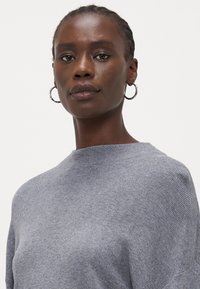 Grå strikkegenser med ribbestruktur, med høy hals og oversized passform. Modellen har på seg sølvfargede hoop-øredobber.