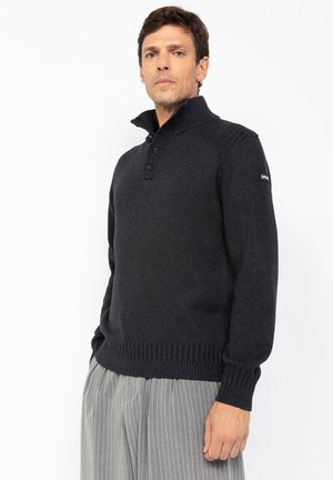 Suéter gris oscuro con un alto cuello, detalles texturizados en los hombros, cierre frontal de tres botones y puños y dobladillo acanalados. Tejido suave, de punto.