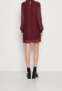 Vestido de encaje burdeos con mangas largas y transparentes y un detalle en forma de ojal en la espalda. El dobladillo es ligeramente transparente, combinado con unos zapatos de tacón grueso negros y calcetines.