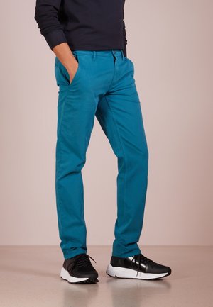 Persoon draagt turquoise strakke broek en zwarte sneakers met witte zolen, staat met handen in zakken tegen een effen achtergrond.