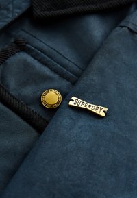 Marineblåt stof med en tekstureret overflade, der har en guldfarvet rund knap og en guldfarvet rektangulær mærkat med teksten "SUPERDRY."