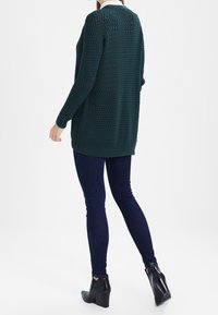 Türkisfarbener gestrickter Pullover mit strukturiertem Design, langen Ärmeln und geripptem Saum, kombiniert mit dunklen Skinny Jeans und Knöchelstiefeln.