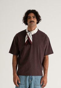 Mand med krøllet hår og overskæg iført en mørkebrun t-shirt, lyseblå jeans og et hvidt mønstret halstørklæde mod en ensfarvet baggrund.