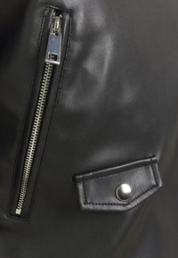 Sezione di indumento in pelle nera con una cerniera verticale in argento e una piccola tasca con pattina dotata di un bottone a pressione in argento.