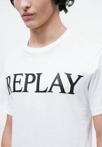 Mlad človek nosi belo majico s poudarjenim črnim napisom "REPLAY" po prsih, skrajšano pri vratu in zgornjih rokavih.