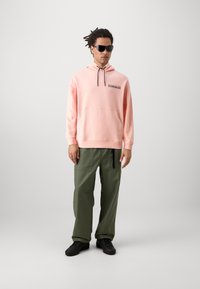 Hoodie rose clair avec cordon de serrage ajustable, poche avant et étiquette imprimée. Assorti avec un pantalon cargo vert olive ample et des chaussures noires.
