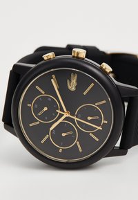 Montre ronde en caoutchouc noir avec accents dorés, cadran noir texturé, trois sous-cadrans pour le jour, la date et l'heure en 24 heures, et détail du logo.
