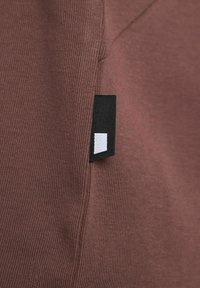 Smeđa rebrasta tkanina s bočnim šavom koji sadrži crnu oznaku i bijeli pravokutni logotip. Usredotočite se na detalje teksture i boje.