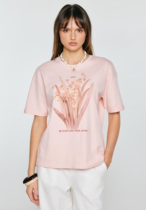 Młoda kobieta ubrana w jasnoróżowy T-shirt w kwiaty z napisem "1979" i "Your Life. Your Jeans.", zestawiona z białymi spodniami i naszyjnikiem z muszelek.