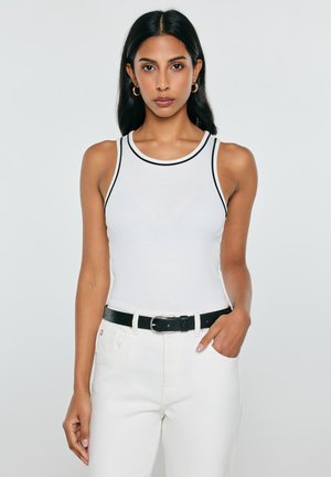 Kobieta z długimi ciemnymi włosami ubrana w biały bezrękawnik z czarną lamówką, białe spodnie i czarny pasek, stojąca na jednolitym jasnym tle.