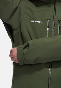 Giacca Mammut verde con colletto alto, tasche con zip e polsini regolabili. Realizzata in materiale impermeabile e dal design elegante.