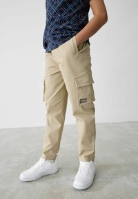 Beige cargobroeken met twee zijzakken en een merklabel; gedragen met witte sneakers en een marineblauw shirt met patroon.