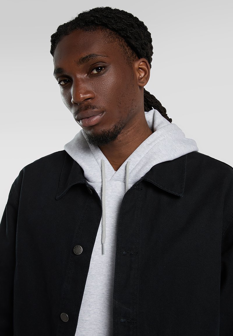 Giacca di jeans nera con felpa grigia con coulisse sotto, con chiusura a bottoni sul davanti e tessuto texturizzato. Primo piano della parte superiore del corpo del modello.